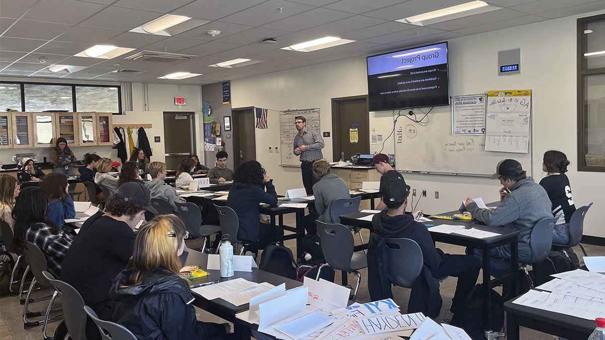 A male teacher stands in front of a classroom of high school students.