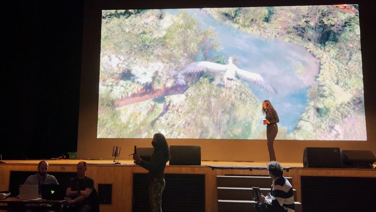 Photo of woman on stage with video playing 和 people in foreground.